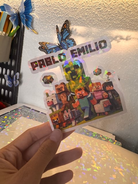 Hand holding a colorful holographic sticker with butterflies and pixel art characters labeled Paolo Emilio